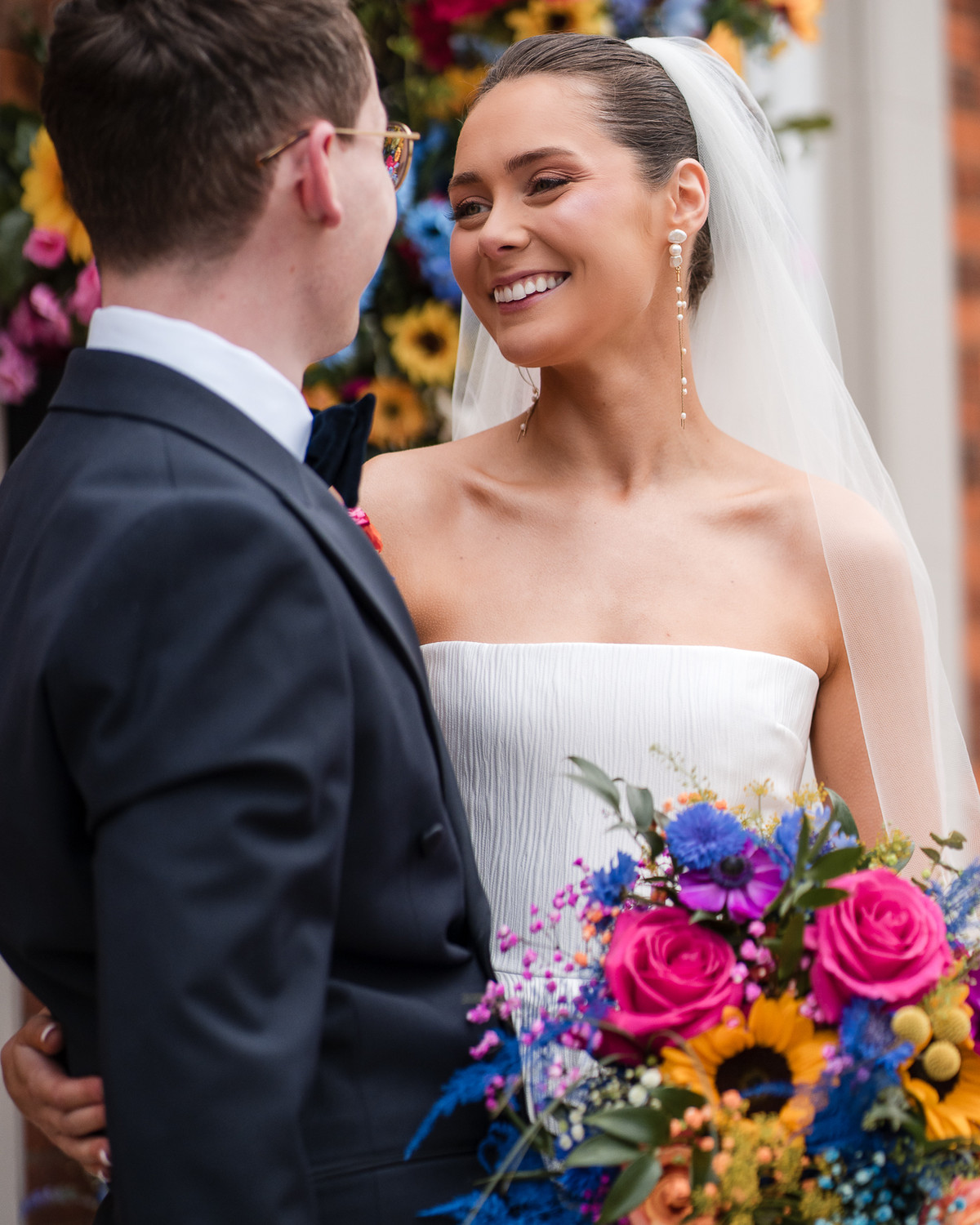 Bride smiling at her groom among colourful summer flowers, by Kevin Joseph Weddings